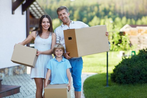 A family of three, consisting of a woman, a man, and a young child, standing outdoors near a residential house in Barnet, with a garden and trees in the background. The woman and man are holding medium-sized cardboard boxes, indicating they are engaged in a house move, with the woman on the left holding a box in her right hand, and the man on the right holding a larger box with both hands. The young child, standing between them, holds a smaller box. The family appears happy and relaxed, dressed in casual clothing suitable for a warm day. The house nearby has a white exterior with a tiled roof, and the setting includes a well-maintained lawn, shrubs, and a paved pathway. This scene reflects the typical activity associated with house removals and local relocation services, showcasing the preparations involved in moving homes within the Barnet area, which is known for its family-friendly residential neighbourhoods. Barnet Man with Van may be implied as the service provider supporting such relocations.
