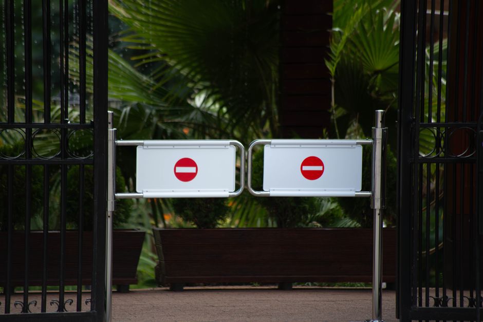 A pair of metal bar barriers with white panels displaying red 'no entry' symbols are positioned across a doorway of a house, blocking access to the outdoor area which is visible behind the gates. The 